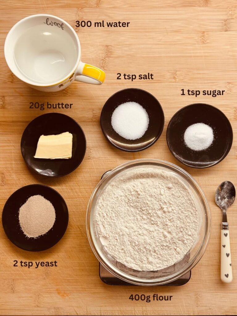 A neatly organized display of bread ingredients on a wooden surface. A labeled cup with 300 ml of water, a bowl with 400g of flour, and small dishes containing 20g of butter, 1 teaspoon of sugar, 1 teaspoon of salt, and 2 teaspoons of yeast