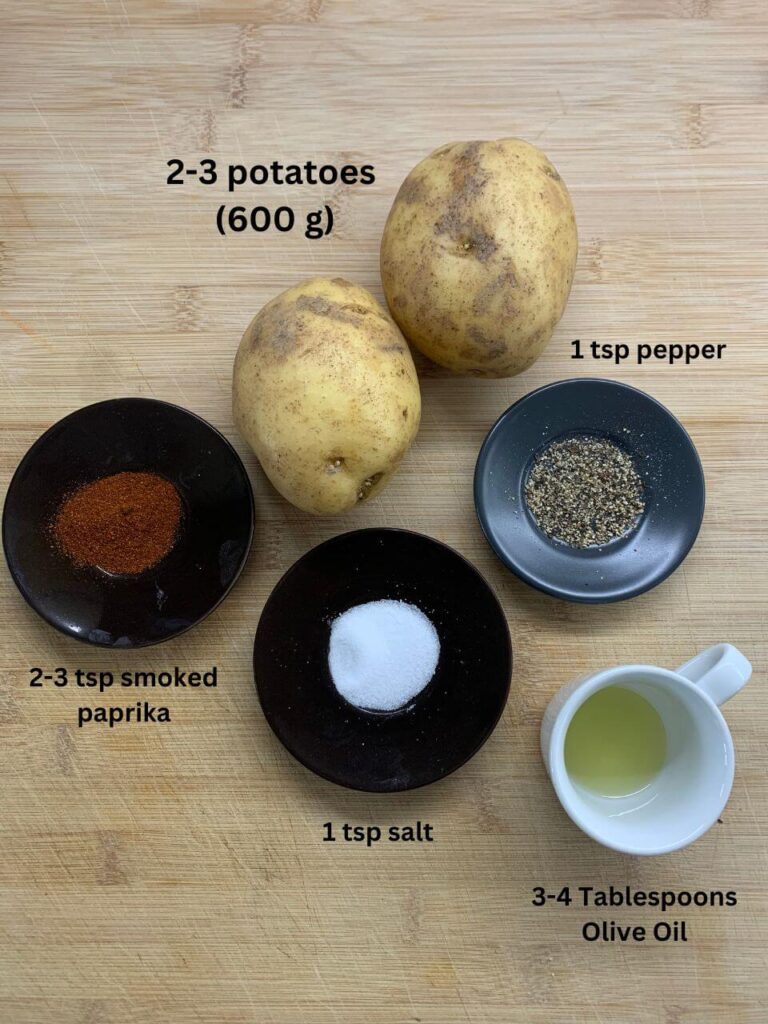 Ingredients for a recipe are displayed: 2-3 potatoes (600 g), 1 tsp pepper, 2-3 tsp smoked paprika, 1 tsp salt, and 3-4 tablespoons of olive oil, arranged neatly on a wooden surface with labels.