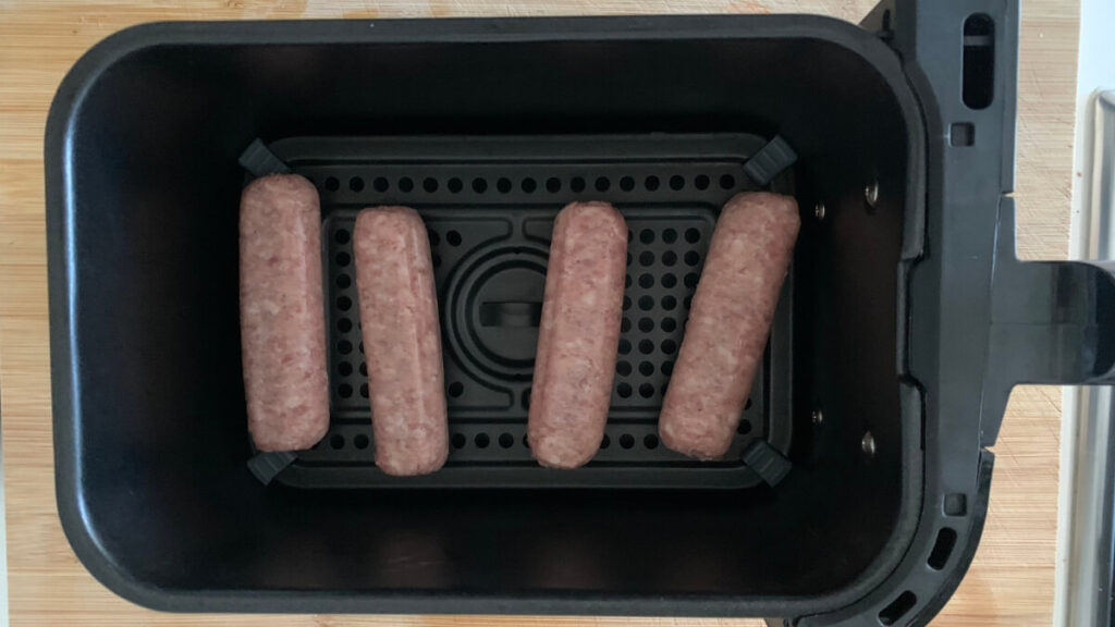 A black air fryer basket with four raw sausages spaced evenly. The sausages are pinkish in color, indicating they are uncooked. They rest on a dark perforated surface with a circular pattern in the middle, and the air fryer handle is visible on the right side.