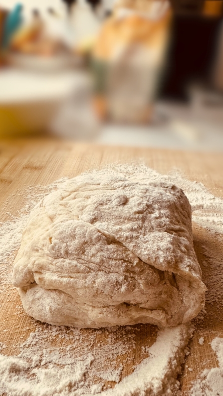 A soft, freshly kneaded loaf of air fryer bread dough rests on a wooden board, blanketed in a fine layer of flour, ready for baking. The background is artfully blurred, drawing focus to the promising start of a delicious bread.