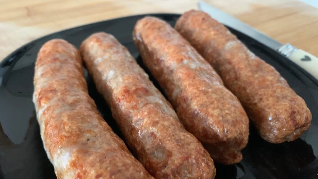 Close-up side angle of four browned sausages on a black plate, showcasing the texture and color more vividly. The cooked sausages have a crispy exterior with visible browning and are arranged neatly in a row.