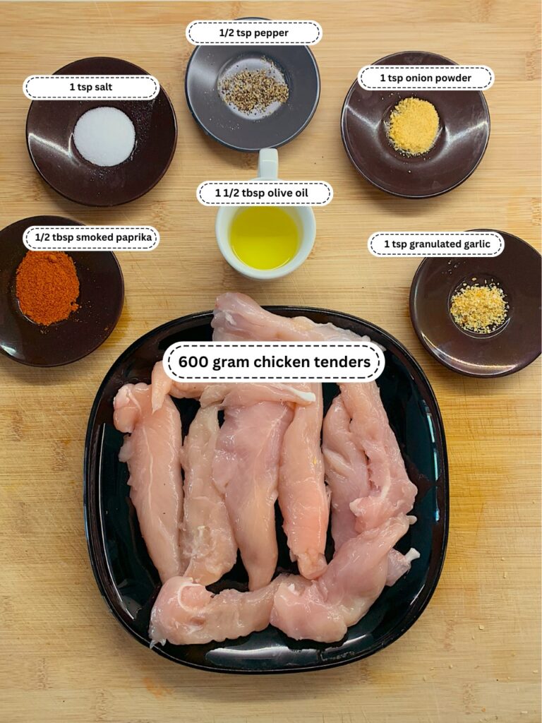 Ingredients for a chicken tenders recipe are neatly organized for preparation. The image shows small dishes containing 1 teaspoon of salt, 1/2 teaspoon of pepper, 1/2 tablespoon of smoked paprika, 1 teaspoon of onion powder, 1 teaspoon of granulated garlic, and 1 1/2 tablespoons of olive oil. A black plate holds 600 grams of raw chicken tenders, and the ingredients are labeled for clarity.