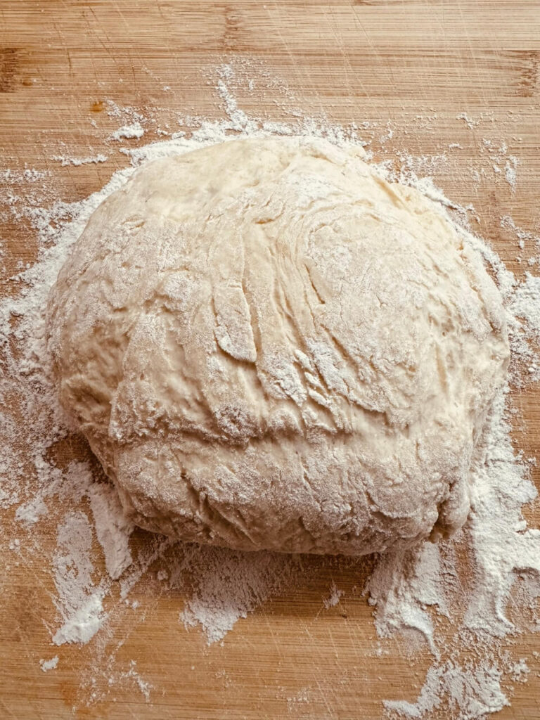 Dough resting on a wooden surface, covered in a light dusting of flour. The dough has a rough texture, indicating that it has been recently kneaded and is now in the midst of the rising process.