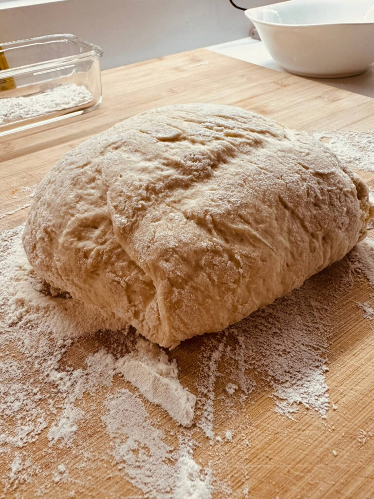 The dough shaped into a round loaf on a floured wooden surface, ready for the next step. The surface shows signs of flour being spread around, indicating the dough has been handled and shaped.
