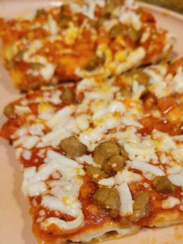 The image captures a cooked Totino's Hamburger Party Pizza on a plate. The pizza has a golden-brown crust with tomato sauce, melted white cheese, and pieces of seasoned meat scattered on top. The pizza is cut into square slices, showing its crispy texture.
