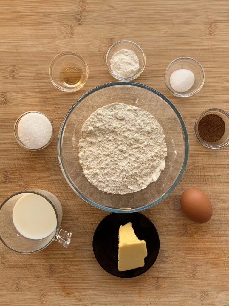 A top-down view of ingredients for homemade donuts on a wooden surface, including bowls of flour, sugar, baking powder, salt, cinnamon, milk, egg, and a small dish of melted butter.