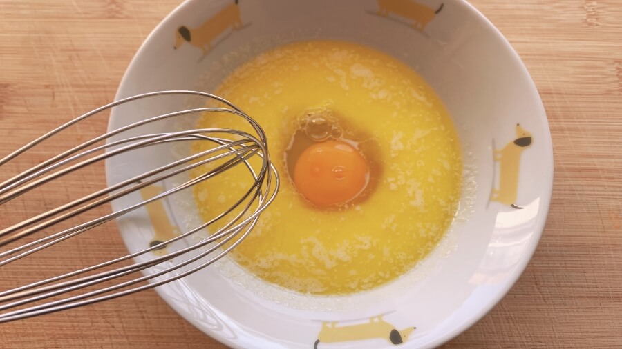 A bowl with a whisked mixture of wet ingredients for donut holes, featuring an egg, melted butter, and vanilla extract in a white bowl with a whisk.