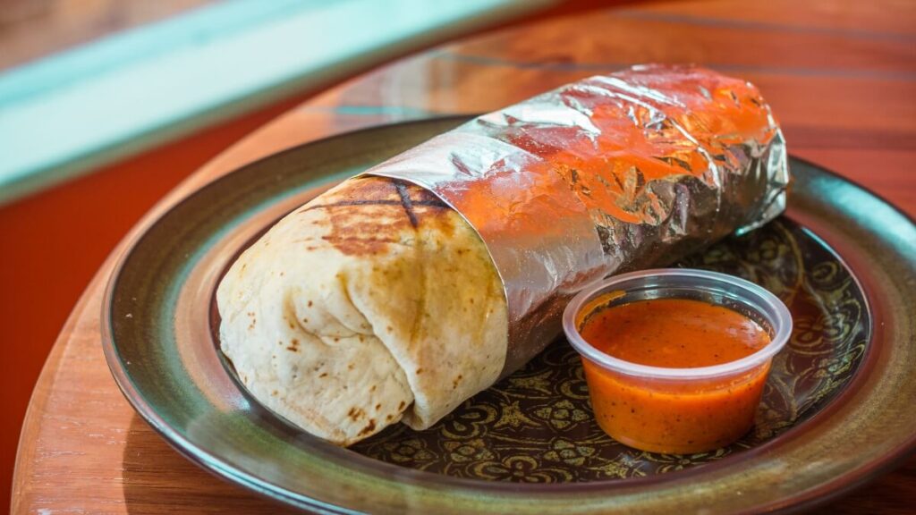 A wrapped burrito with visible grill marks on the tortilla, secured in foil, placed on a decorative plate next to a small plastic container of red salsa.