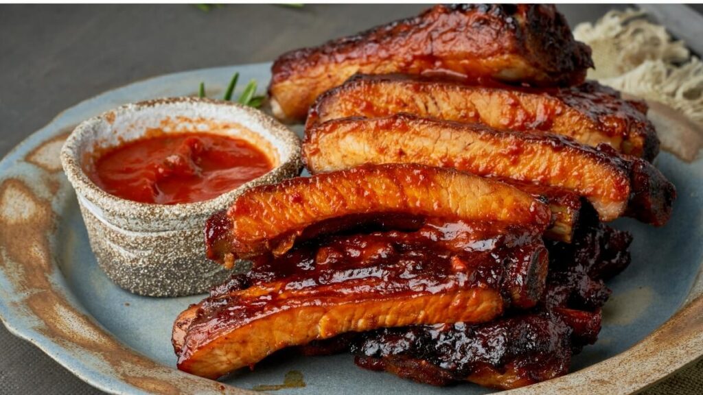Smoky barbecued pork ribs with a caramelized glaze, served on a ceramic plate with a side of red sauce in a rustic bowl, garnished with a sprig of rosemary.