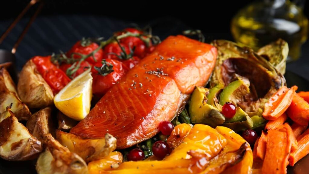 A colorful plate of roasted foods including a glazed salmon fillet, cherry tomatoes on the vine, lemon wedges, and a variety of roasted vegetables such as carrots, bell peppers, and potatoes, all garnished with herbs.